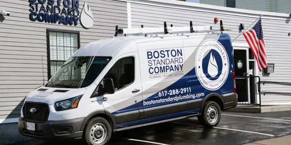 A Boston Standard Company plumbing van is parked outside their office, ready for its next call. Nearby, an American flag waves proudly. This trusted team is known for top-notch service, including expert heat pump installations.