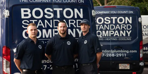 Three men in uniform stand in front of two vans displaying the "Boston Standard Company" logos, ready to tackle any heating issue, specializing in heat pump maintenance.