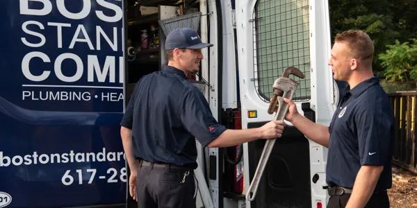 Two workers in uniforms stand next to a van, one handing a large wrench to the other. The van is emblazoned with branding related to plumbing and heating, specializing in burst pipe repair.