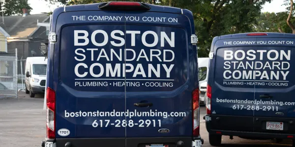 Rear view of vans with "Boston Standard Company" signage, highlighting expertise in plumbing, heating, cooling, and tankless water heater maintenance services, complete with contact details.