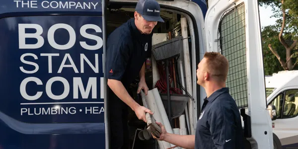 Two workers in matching uniforms unload equipment from a company van labeled "Boston Standard Co." While handing tools to each other, they're preparing for a tankless water heater repair.