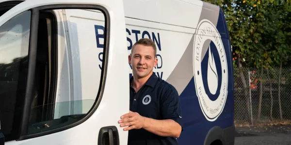 A person stands by the open door of a white and blue van with "BOSTON SEWER & DRAIN" emblazoned on the side, ready for the day’s tasks, including essential water heater maintenance.