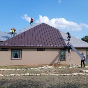 Four Roofing contractors installing a dark red metal roof on a tan house under a sunny sky in San Antonio, TX