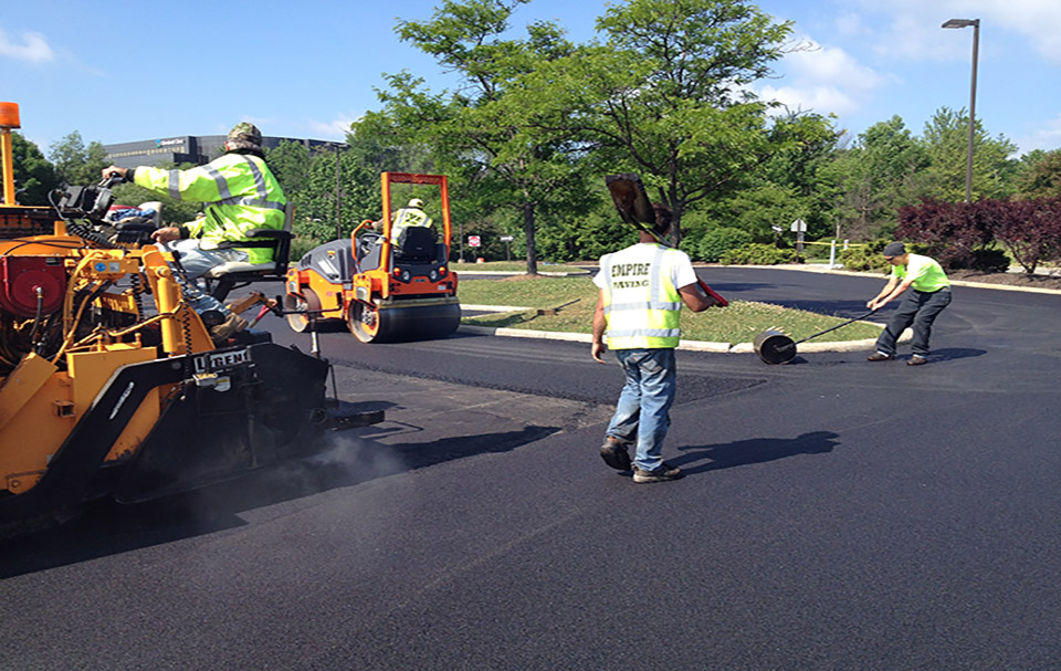 Three construction workers pave a road using heavy machinery, including an asphalt paver and steamroller, on a sunny day. As a local Asphalt Paving Company, they expertly handle equipment while trees and greenery form the background.