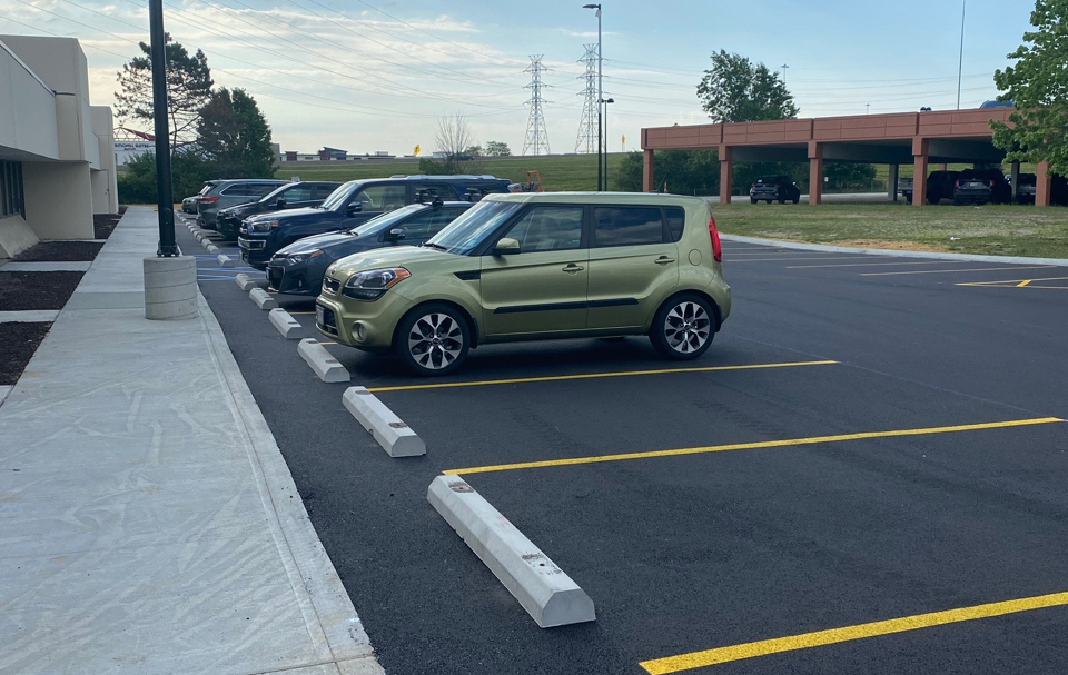 A small green car is parked in an almost empty parking lot, recently finished by a local paving contractor. The sky is partly cloudy, and there are trees and a grassy area in the background.