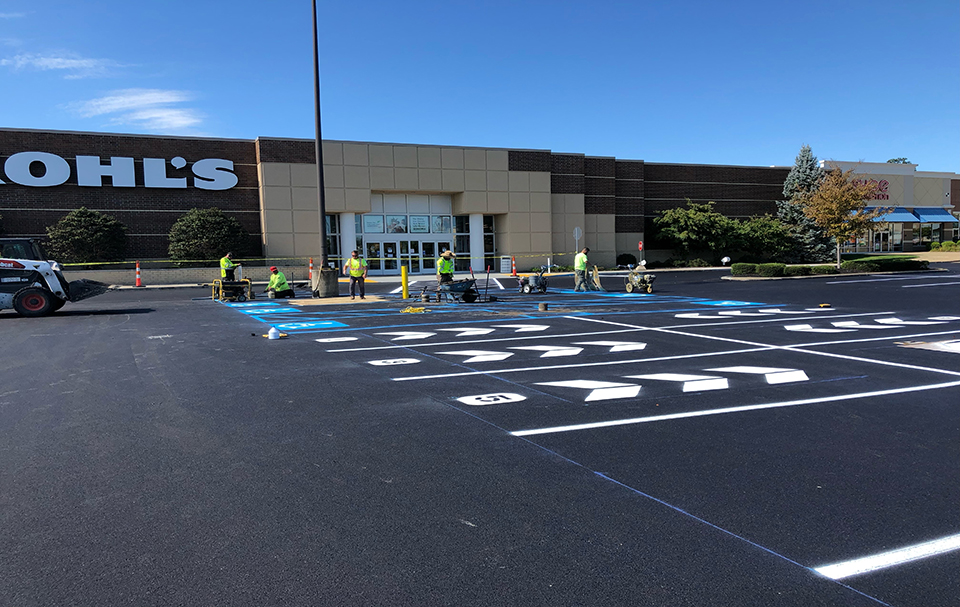 Workers in safety vests paint lines and symbols on a freshly paved parking lot by an Asphalt Paving Company in front of a Kohl’s store on a clear, sunny day. Parking spaces and directional arrows are visible in the foreground.