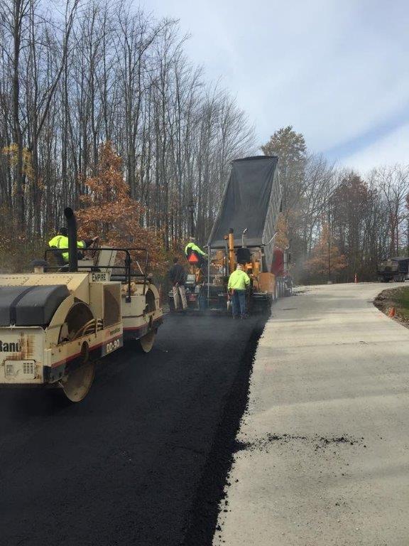 Construction workers from an Asphalt Paving Company operate heavy machinery as they lay fresh asphalt on a road. A roller compacts the asphalt while another machine spreads it. Trees with autumn leaves line the background.