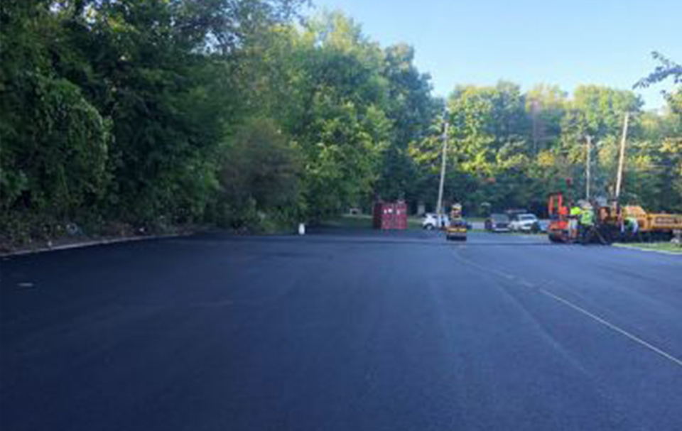 Freshly paved parking lot by a skilled asphalt contractor, with construction equipment and several workers near the far edge, surrounded by green trees and parked cars in the background.