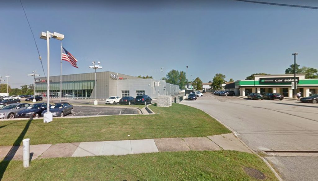 A car dealership lot with parked cars on freshly paved asphalt, an American flag on a pole, a modern gray showroom building, and a green sign for car sales on a separate building, all enhanced by expert asphalt installation on a sunny day.