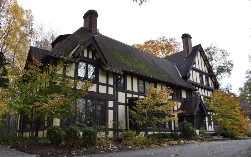 A large Tudor-style house with tall chimneys, dark wooden beams, cream-colored walls, and diamond-paned windows sits among autumn trees and a paved driveway installed by a trusted paving contractor.