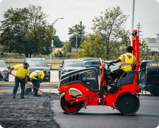 Three workers in safety vests repave a parking lot; two use rakes for asphalt installation while the third operates an orange road roller. Parked cars and trees are visible in the background, highlighting the expertise of this paving contractor.