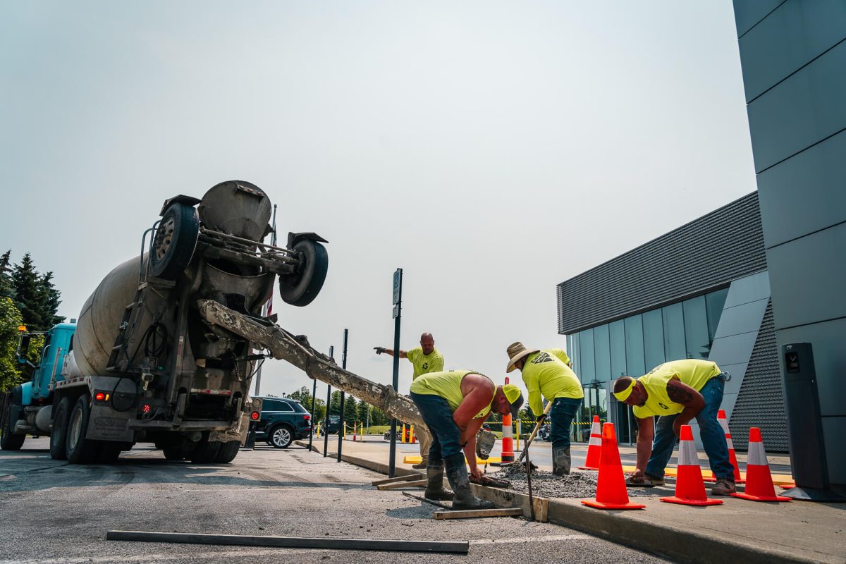 Four construction workers in yellow shirts pour and spread concrete from a mixer truck onto a sidewalk, surrounded by orange traffic cones near a modern building—a professional paving contractor at work.