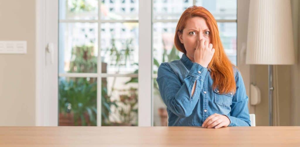 A person with red hair sits at a table, holding their nose and making a displeased expression. They are wearing a blue shirt, and a window is visible in the background.