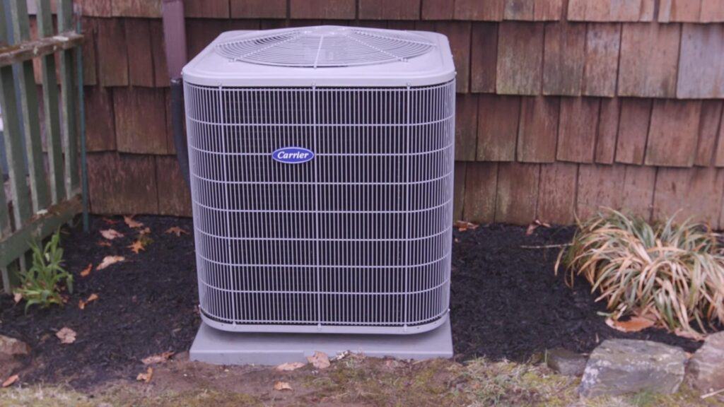 A gray outdoor air conditioning unit discreetly sits next to a wooden wall, surrounded by mulch and grass. Its sleek design blends seamlessly with the environment, while the hidden warranty ensures lasting peace of mind.