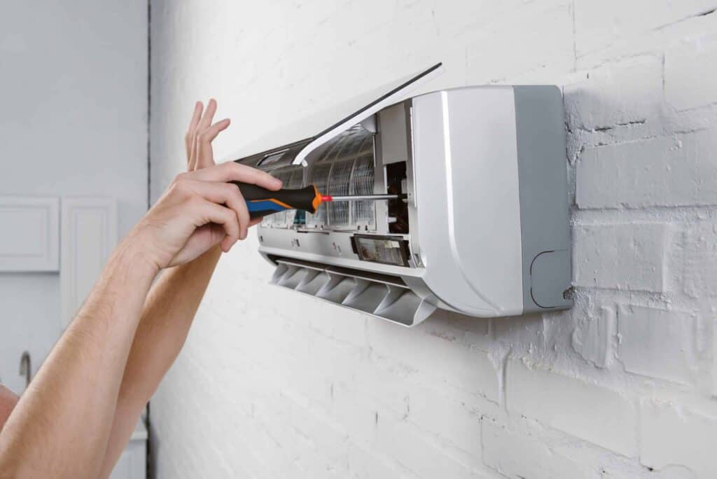 Person using a screwdriver to fix a frozen A/C line on an air conditioning unit mounted on a white brick wall.