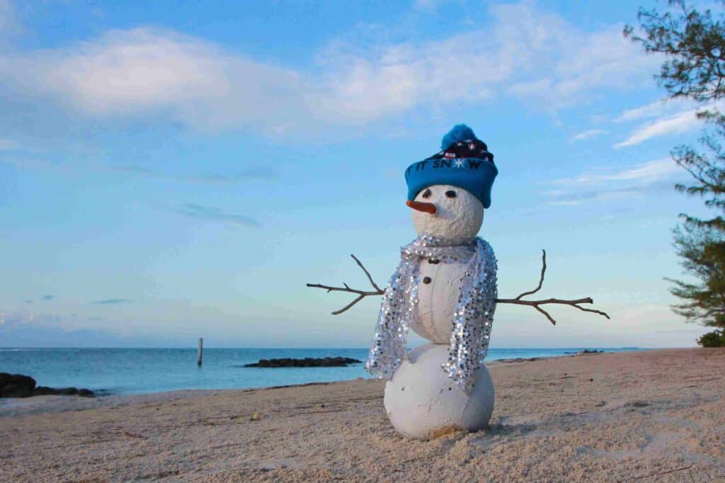 A snowman with a blue hat and scarf stands on a sandy beach near the ocean under a bright blue sky, offering an unexpected scene as you consider winter preparation for your Florida home plumbing.