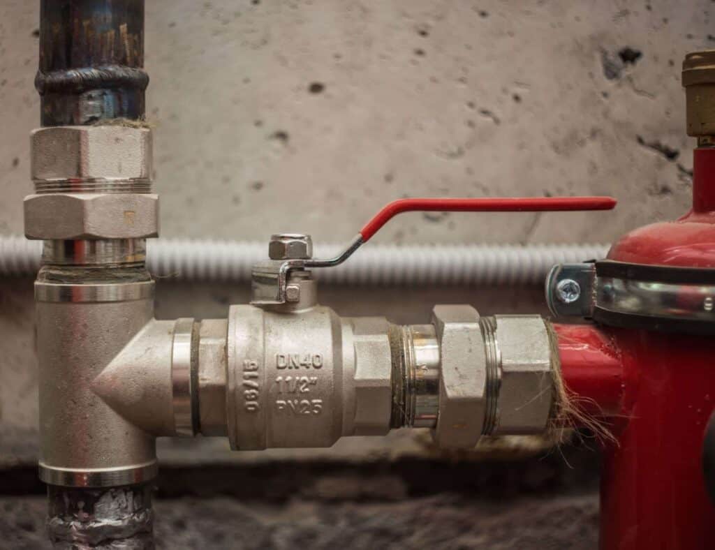 A metal pipe system with a red-handled main water shut off is connected to a red cylinder against a concrete wall.