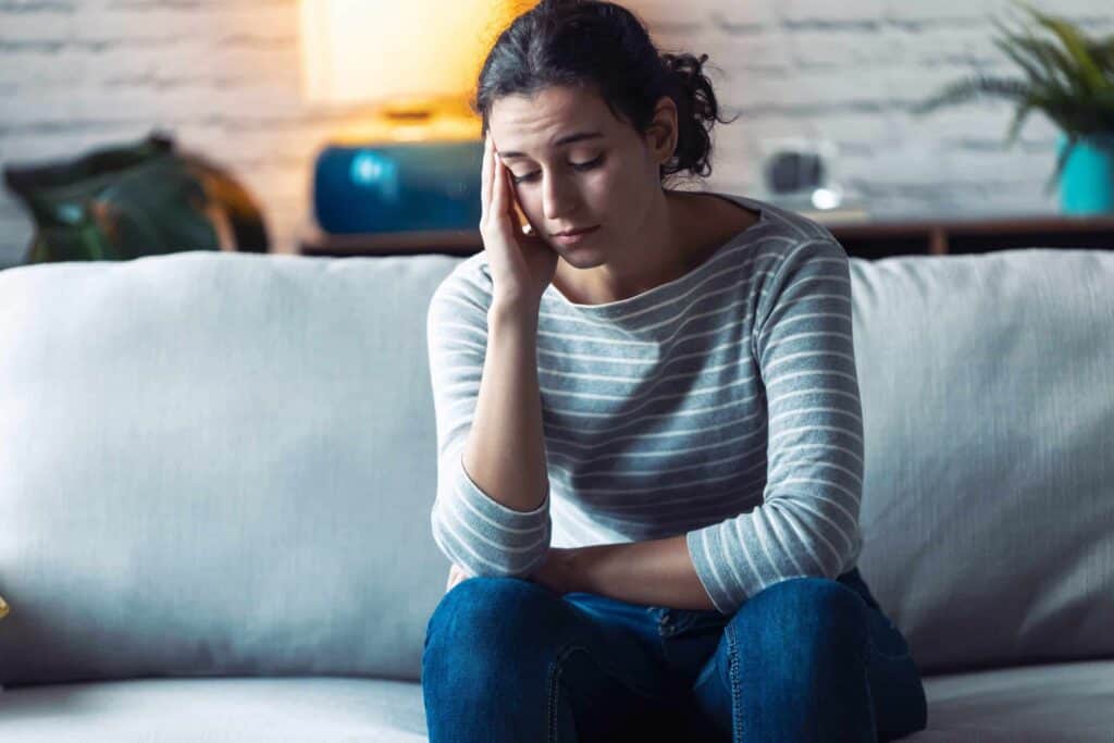 A person sits on a couch with one hand on their face, appearing thoughtful or tired. They wear a striped shirt and jeans, perhaps contemplating the AC issues that have been keeping them up at night. A lamp and decor are visible in the background, casting a soft glow over their pensive expression.