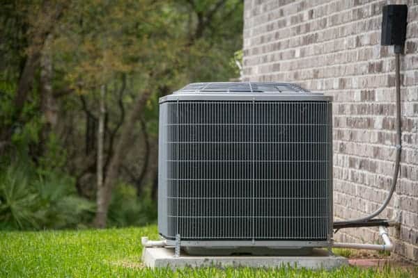 Outdoor air conditioning unit beside a brick wall, situated on a concrete slab surrounded by grass.