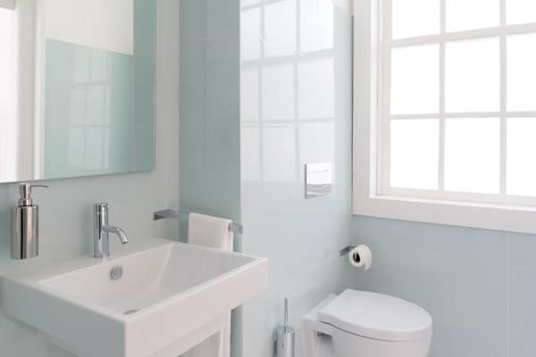 A small, clean bathroom with a white sink, a wall-mounted faucet, a soap dispenser, a toilet, and a towel holder. A large window on the right allows natural light in.