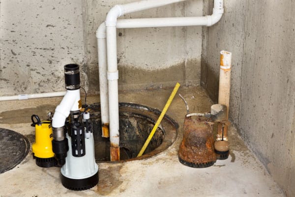 A basement sump pit with white PVC pipes and multiple sump pumps, surrounded by concrete walls and a damp floor.