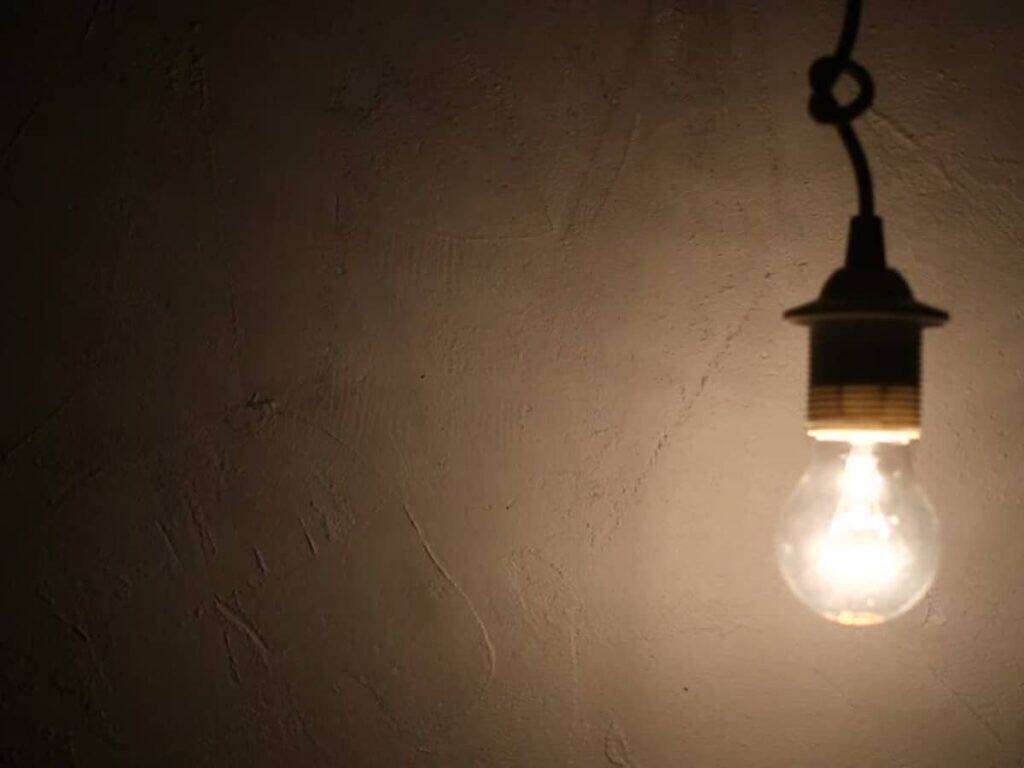 A single lit light bulb hangs from the ceiling, casting a dim glow on a textured wall, as if flickering lights hinted at underlying electrical issues.