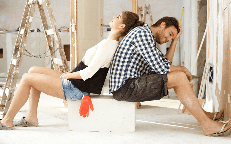 A man and woman, exhausted from DIY home projects, sit back-to-back on a box in a room under renovation. Ladders and construction materials surround them.