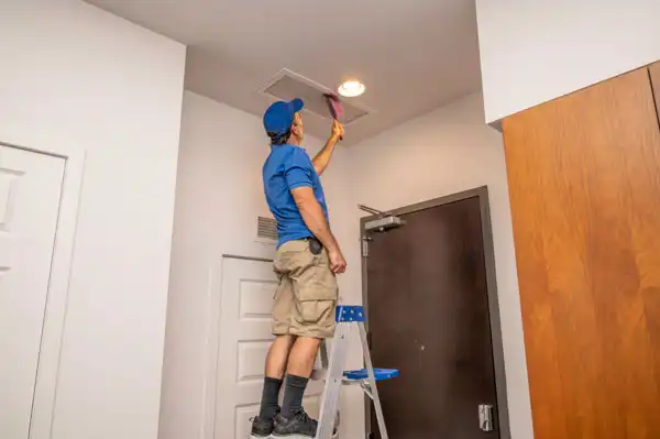 A person wearing a blue shirt and beige shorts stands on a small ladder, dusting a ceiling vent with a pink duster in a room with white walls and a wooden door.