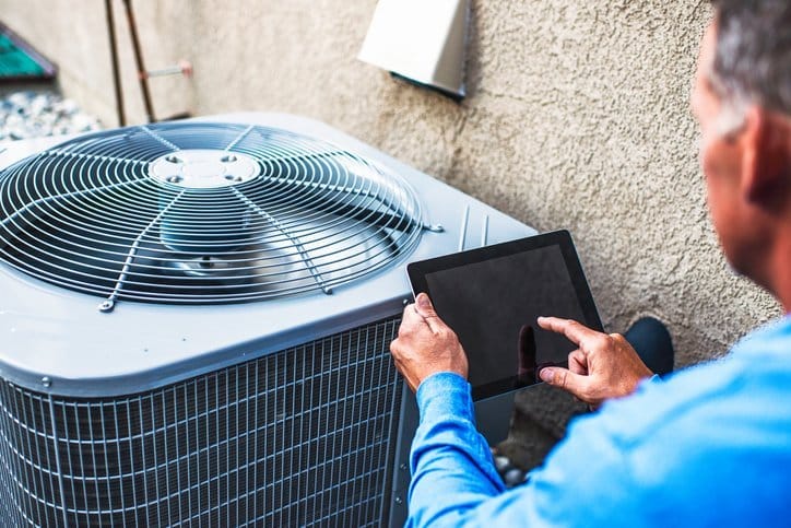 A person in blue shirt uses a tablet to inspect an outdoor air conditioning unit.