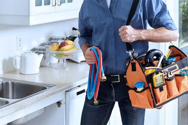 A person holding a tool bag and colored hoses stands in a kitchen near the sink, ready for plumbing repair. A fruit bowl and tea kettle are visible on the countertop, creating a cozy yet functional atmosphere.