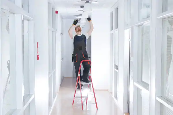 Person on a ladder in a hallway, wearing a black apron and gloves, adjusting an overhead light fixture.