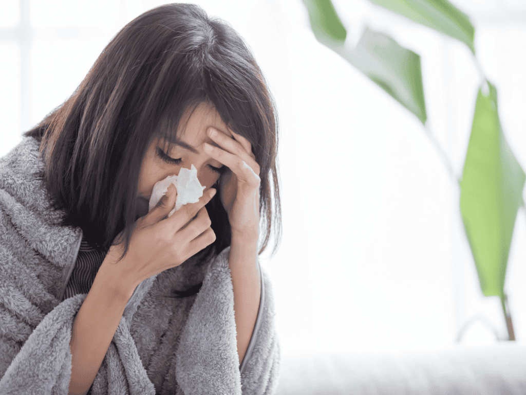 A woman with a blanket blowing her nose with a tissue, while holding her head.