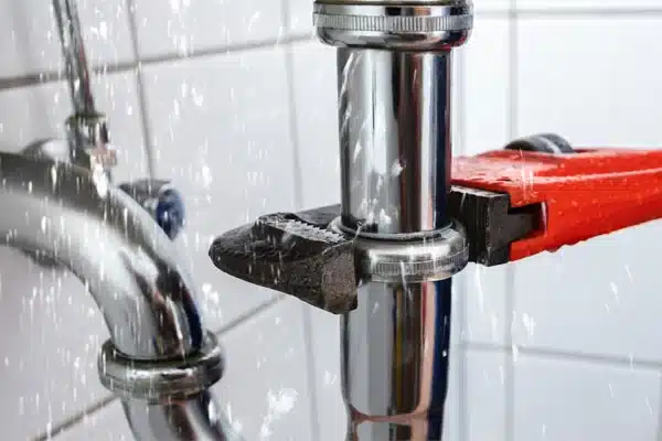A red pipe wrench is tightened around a leaking metal pipe joint in a tiled setting, with water spraying out of the connection.
