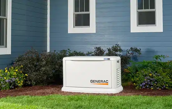 Generac home standby generator installed next to a blue house with two windows, surrounded by shrubs and flowers, ensures reliable backup power. Regular home generator maintenance is essential to keep everything running smoothly.