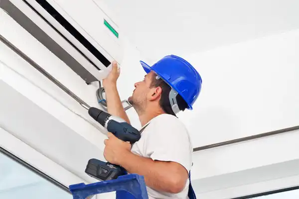 A technician in a blue hard hat standing on a ladder installs an air conditioning unit, holding a power drill.