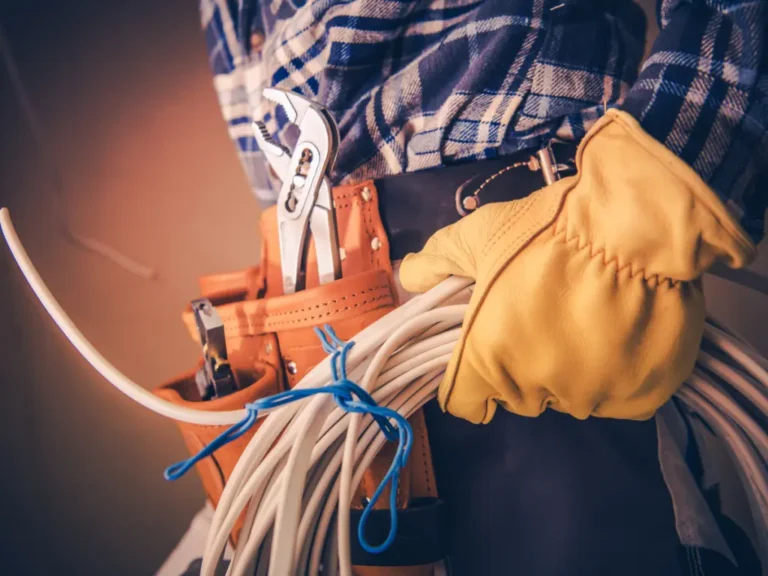 Person wearing gloves and a plaid shirt, holding coiled cables with a tool belt carrying pliers and other tools.