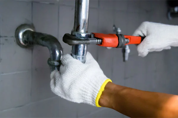 Person wearing gloves uses a wrench to tighten a pipe against a tiled wall.