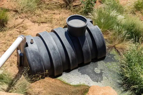 A black plastic septic tank is partially buried in a grassy area, with a pipe connected to it. Some water and algae are visible around the tank, hinting at the need for septic tank repair to ensure everything flows smoothly.