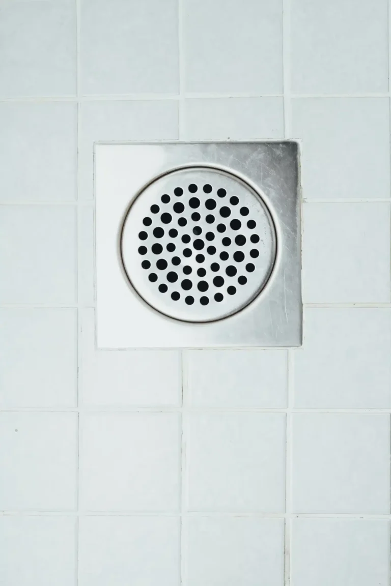 Square metal drain cover with circular perforations on a tiled surface, perfect for ensuring efficient drainage. For reliable maintenance or Rooter Services in Rockville MD to keep it clear and functional, trust local experts to handle your plumbing needs.