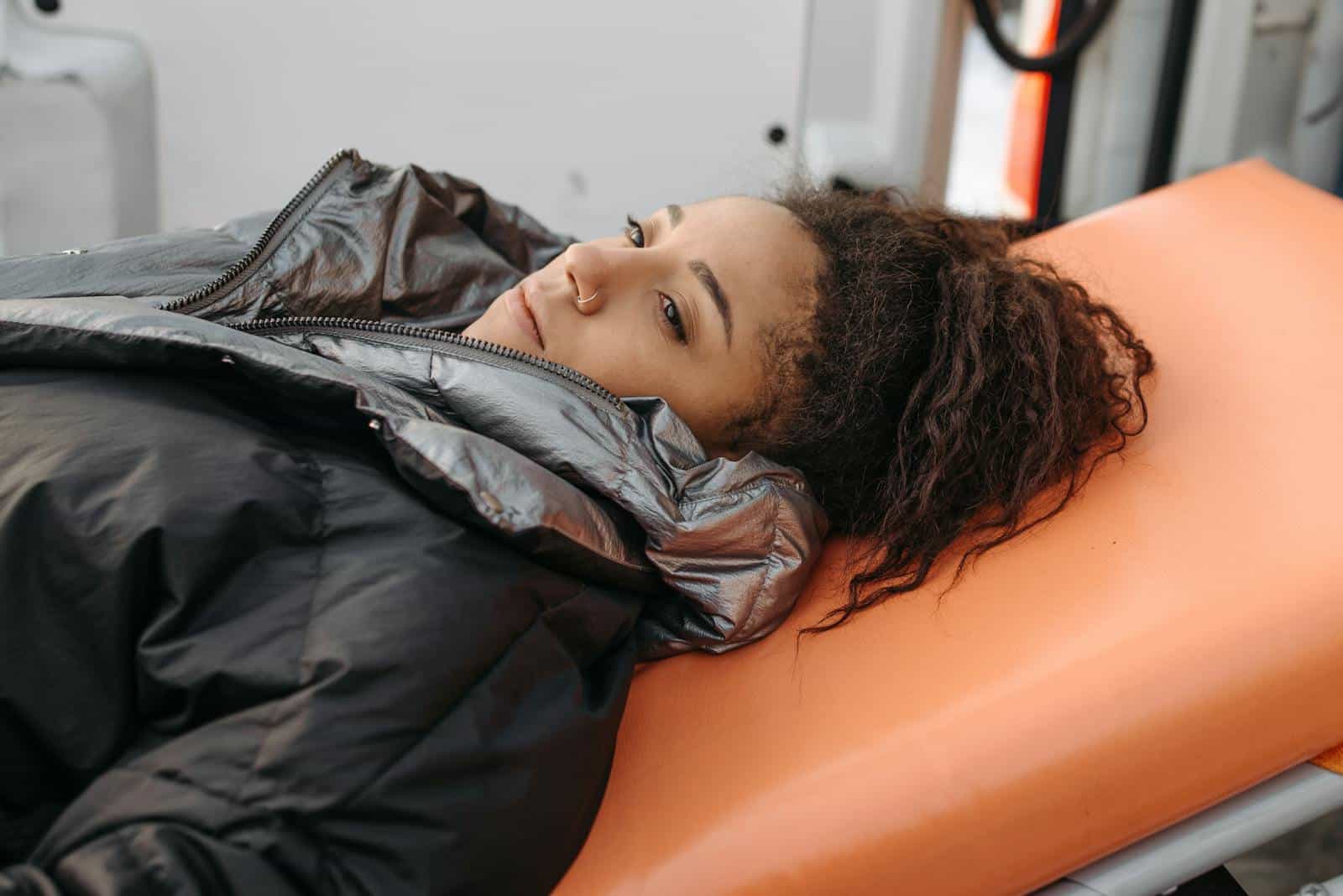 A woman in a padded jacket lies on a stretcher, appearing contemplative and unwell.