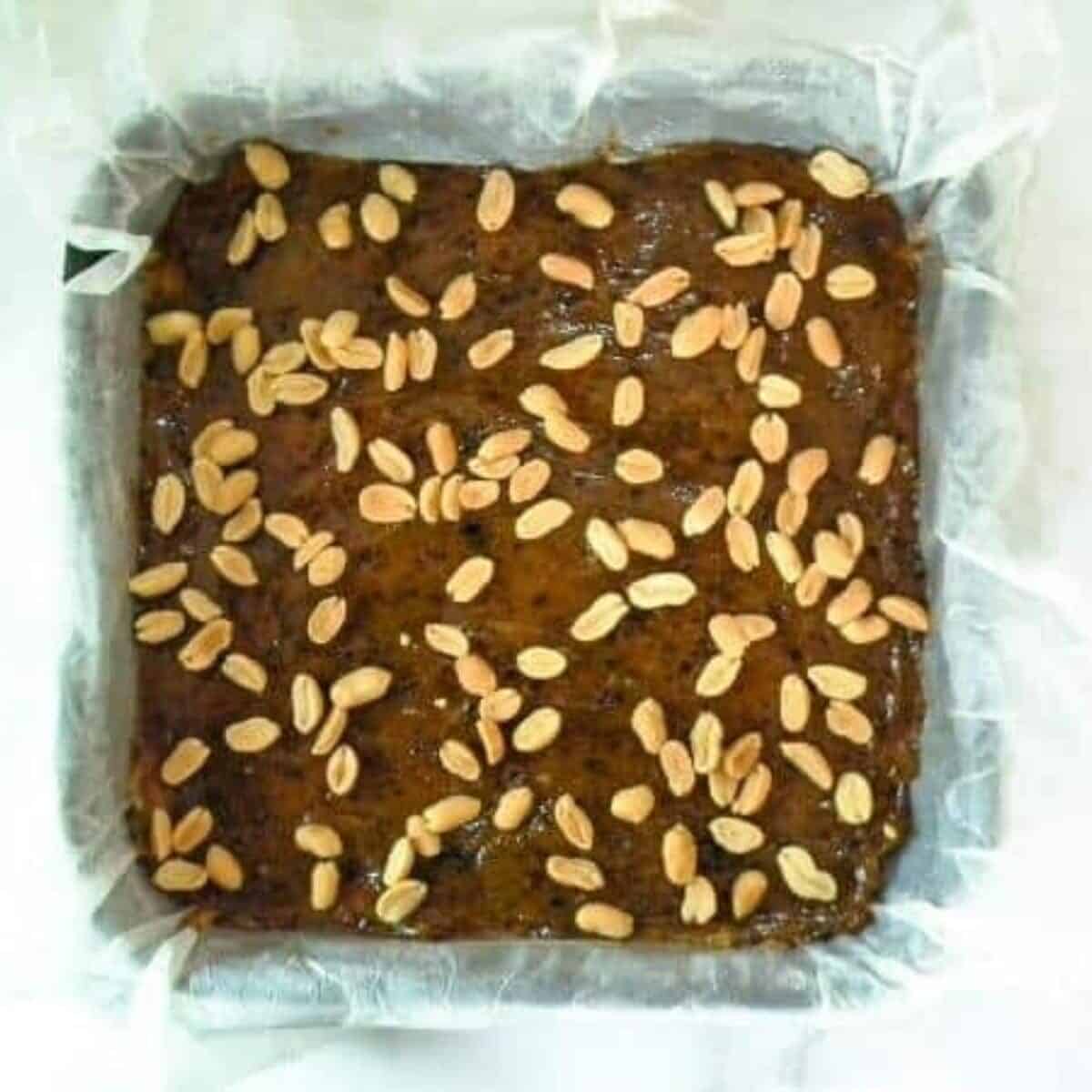 A square cake reminiscent of homemade Snickers bars, with scattered peanuts on top, rests in a parchment-lined baking dish.