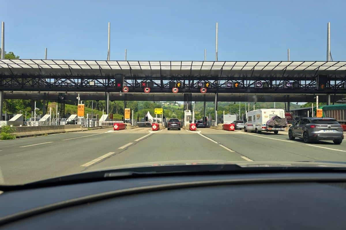 As you drive in France, the view from your car approaching a highway toll booth reveals several open lanes. Traffic signs and lights above indicate lane information, while vehicles are visible ahead and to the right under the clear sky.