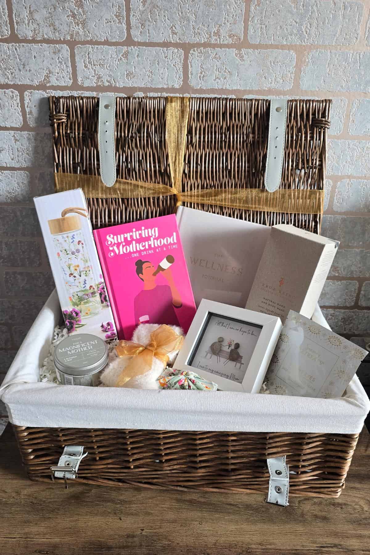 Wicker gift basket thoughtfully curated for Mother's Day, featuring books, a photo frame, candles, chocolates, and wellness items—all set against a rustic brick wall. It's the perfect choice from any gift guide to show your appreciation for mum.