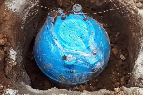 A blue plastic water tank wrapped in protective film is partially buried in a dirt pit, prepared for septic tank installation in Fairfax, VA.