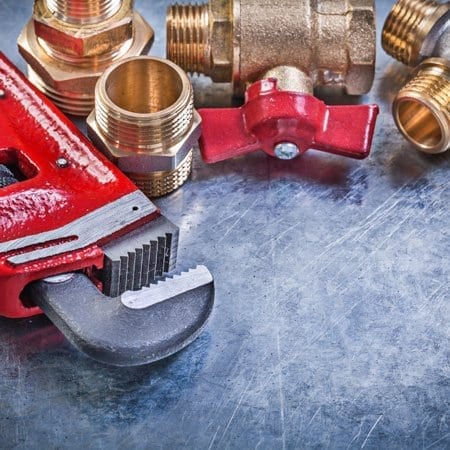A red pipe wrench and various brass plumbing fittings, including a valve and couplers, are arranged on a scratched metal surface.