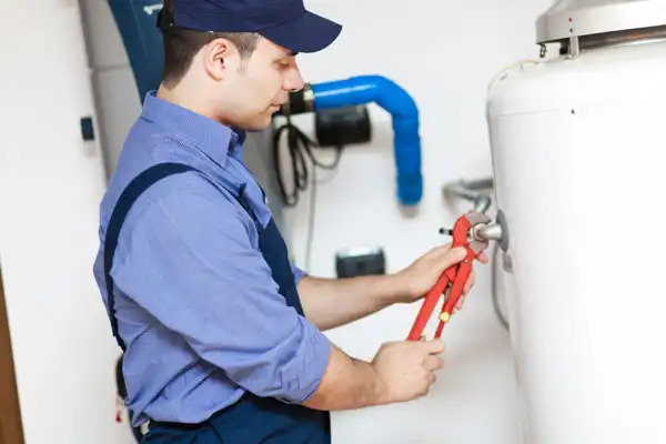 A plumber in a blue uniform and cap performs water heater maintenance with a wrench on a tank in a Van Nuys, CA utility room.