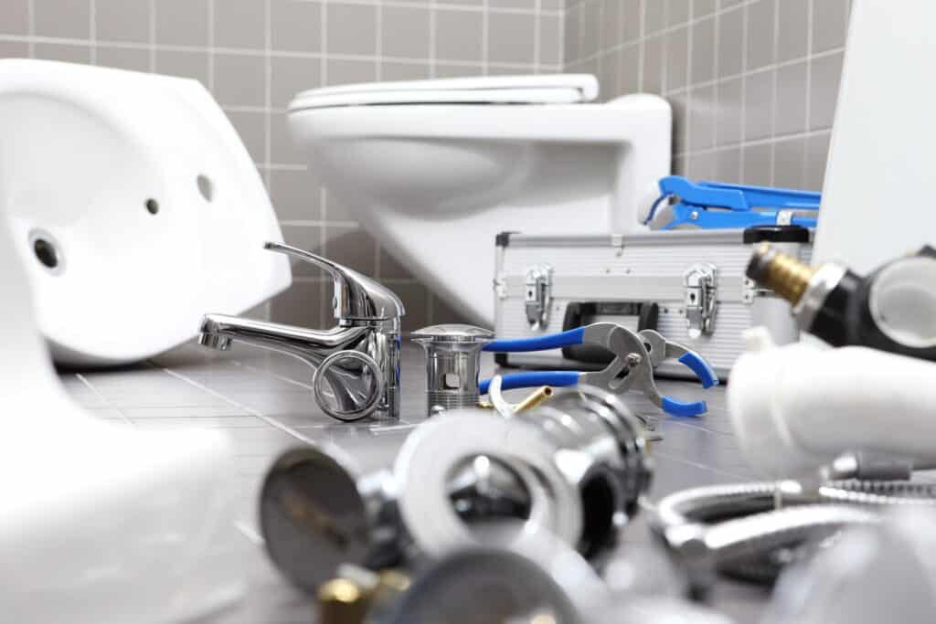Plumbing tools and fixtures are spread out on a tiled bathroom floor near a toilet and a toolbox, indicating ongoing repair or installation work.