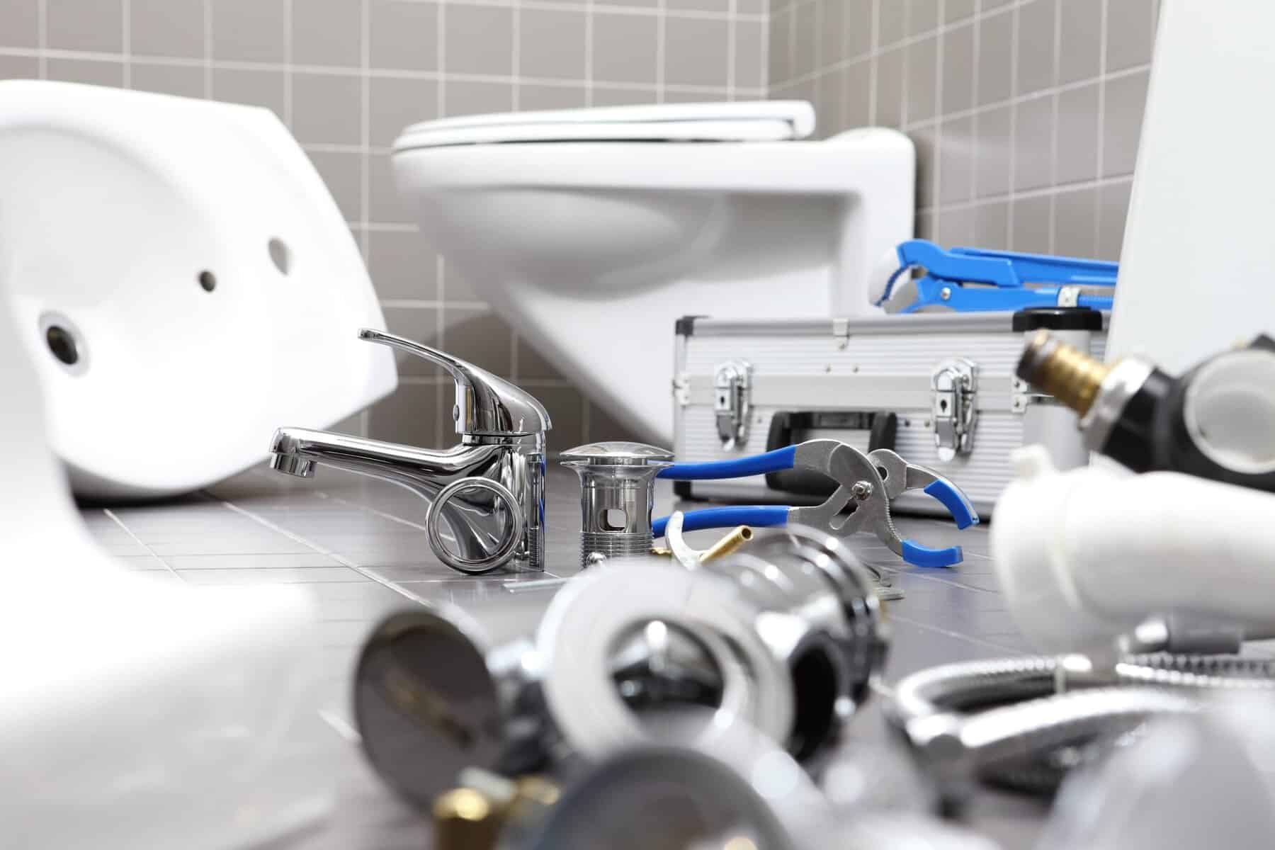 Plumbing tools and fixtures are spread out on a tiled bathroom floor near a toilet and a toolbox, indicating ongoing repair or installation work.