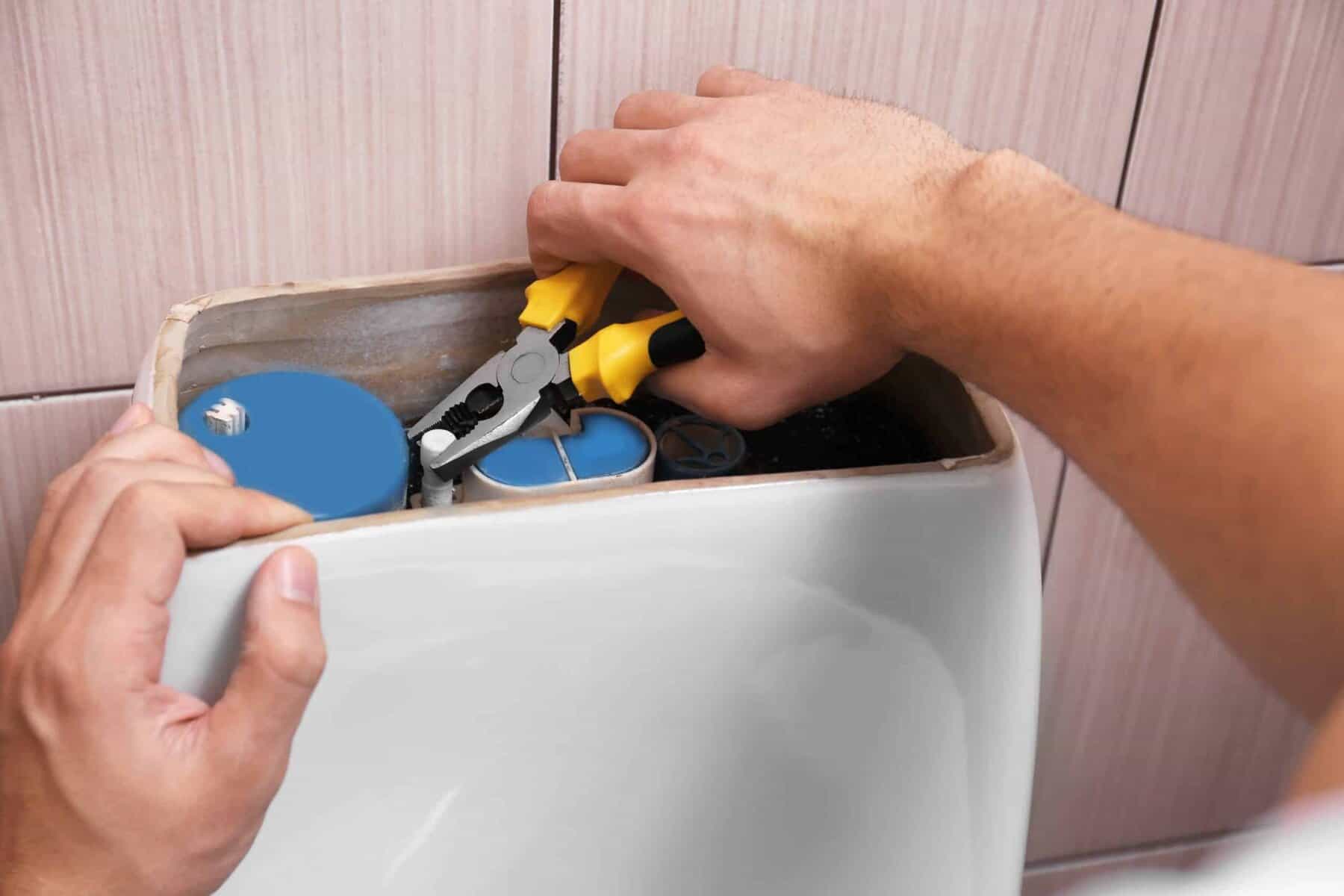 A person uses pliers to repair the internal mechanism of a toilet tank against a tiled wall.