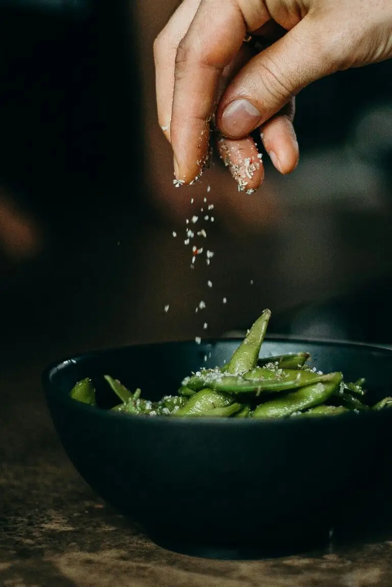 a hand salt sprinkling on edamame beans in a black bowl